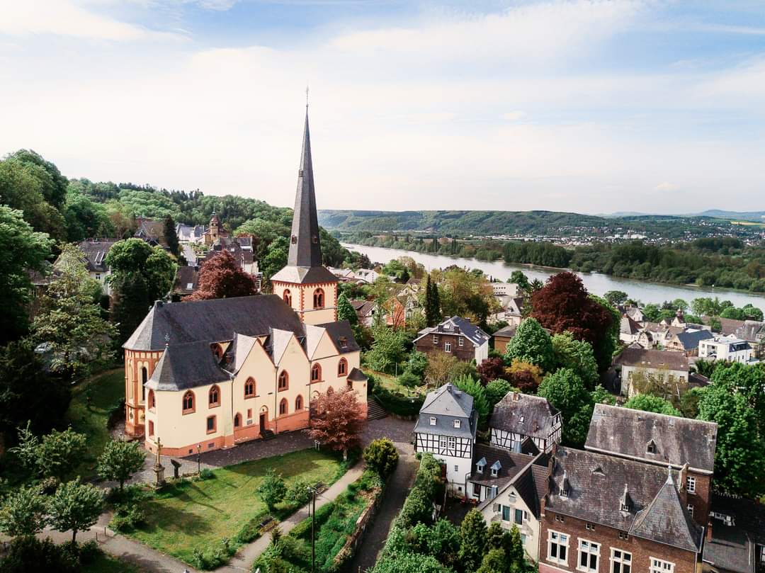 Luftbild der Kirche St. Martin in Linz am Rhein mit Fachwerkhäusern und Kirchgarten im Vordergrund und dem Rhein und der Eifel im Hintergrund.